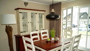 Uma sala de jantar com uma mesa de madeira, cadeiras brancas, um armário com portas de vidro, um lustre e vista para o exterior através de grandes janelas.