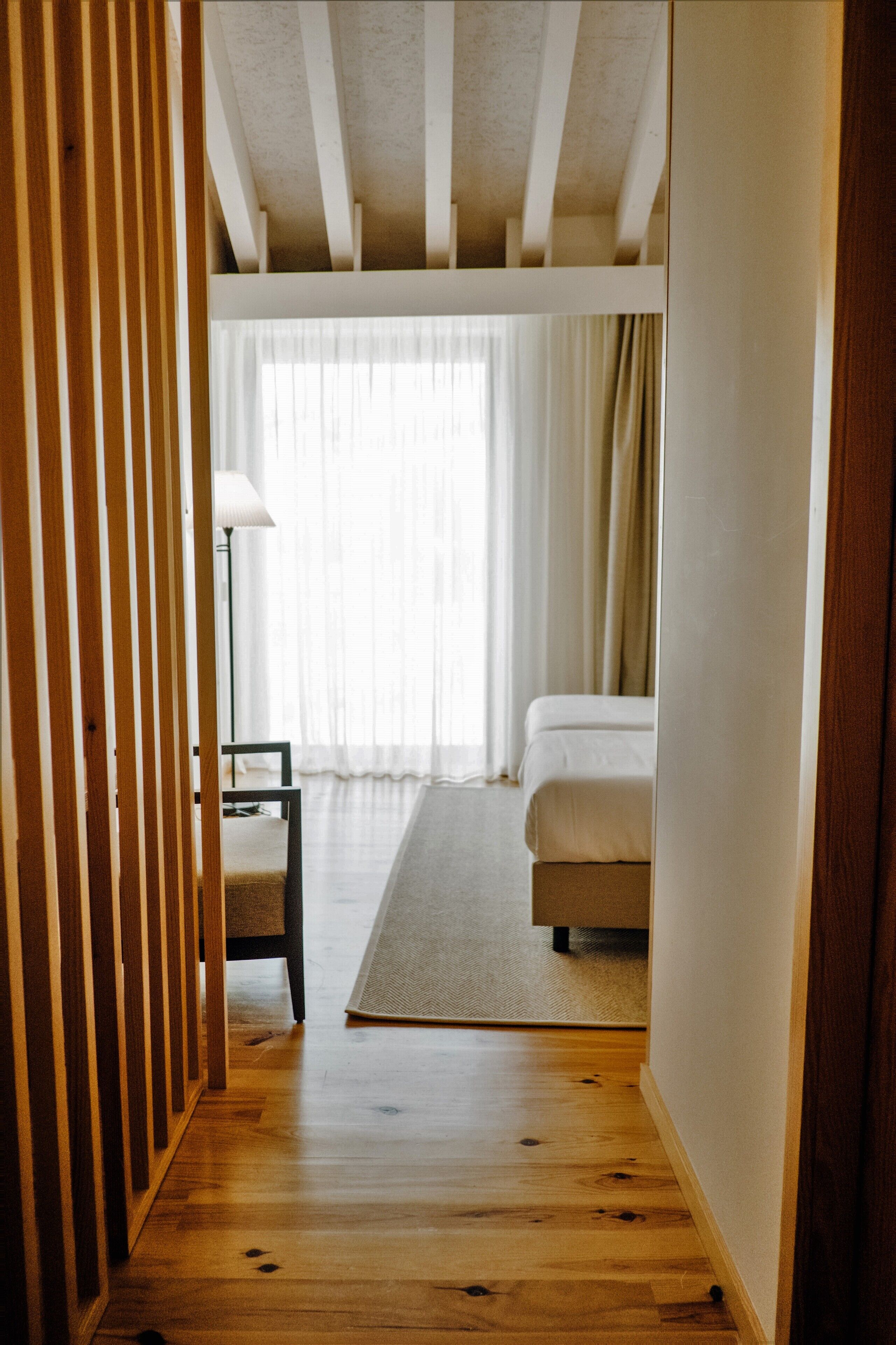 A narrow hallway leading to a bedroom with a bed, a chair, and a lamp.