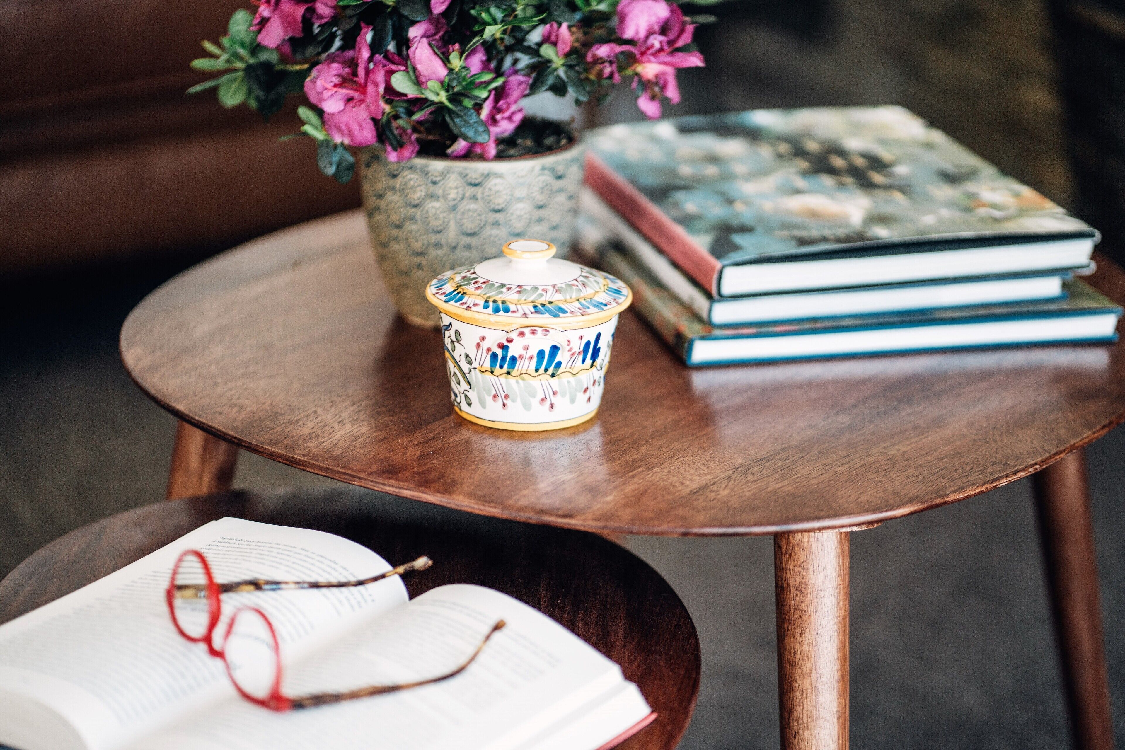 A round wooden table with a ceramic container, a potted plant with pink flowers, and a stack of books.