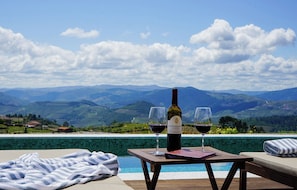 Escena junto a la piscina con una botella de vino, copas, una toalla y vistas a montañas distantes.