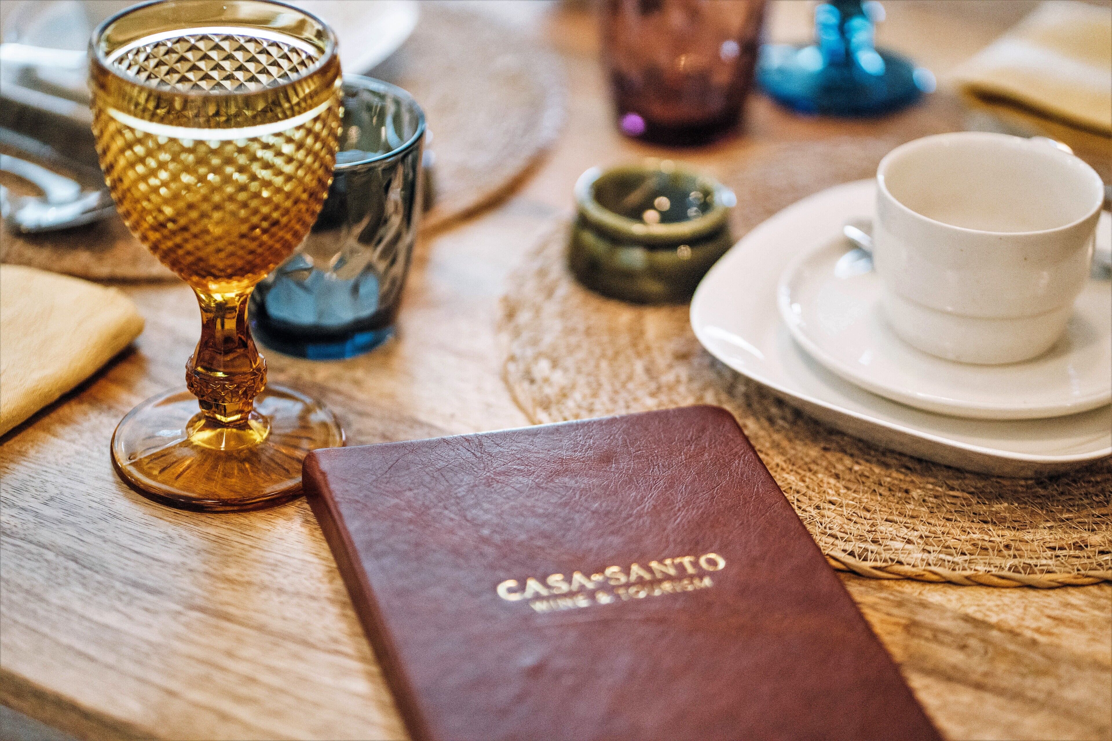 Uma mesa com um livro intitulado 'CASA SANTO WINE & TOURISM', um copo com textura e uma chávena branca sobre um pires.