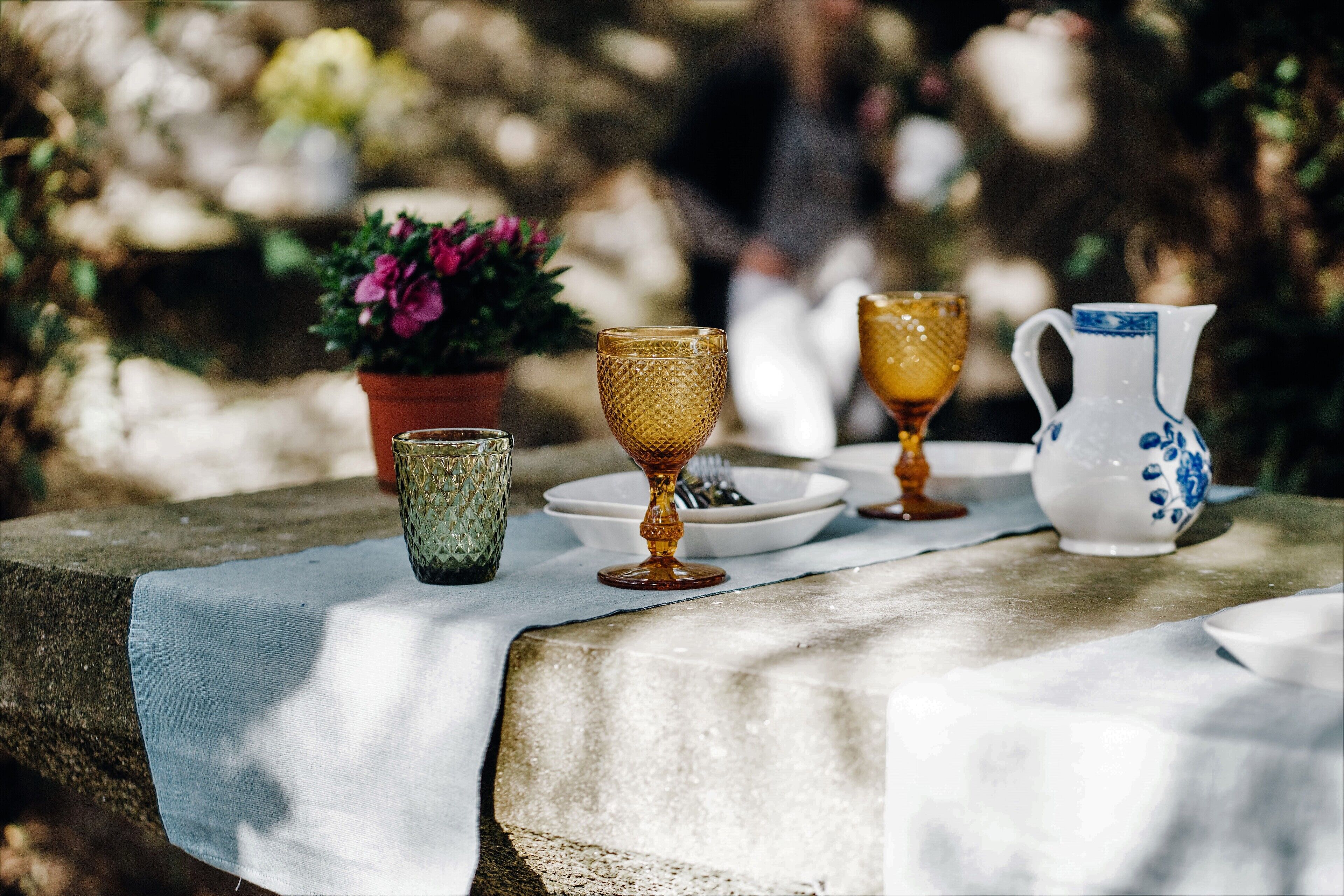 A table with a white cloth, a green glass, a golden goblet, a yellow glass, and a white pitcher with blue designs.