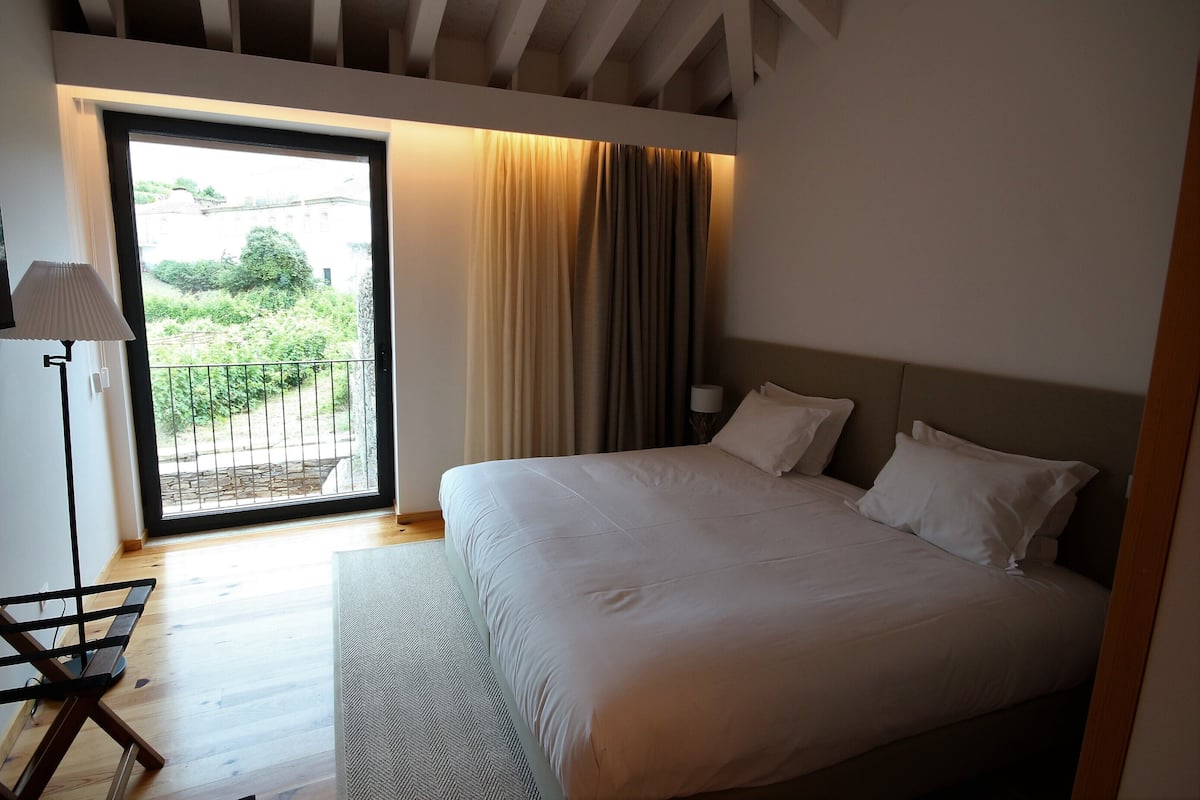 A bedroom with a large bed, a nightstand with a lamp, a view of greenery through a glass door, and a wooden floor.