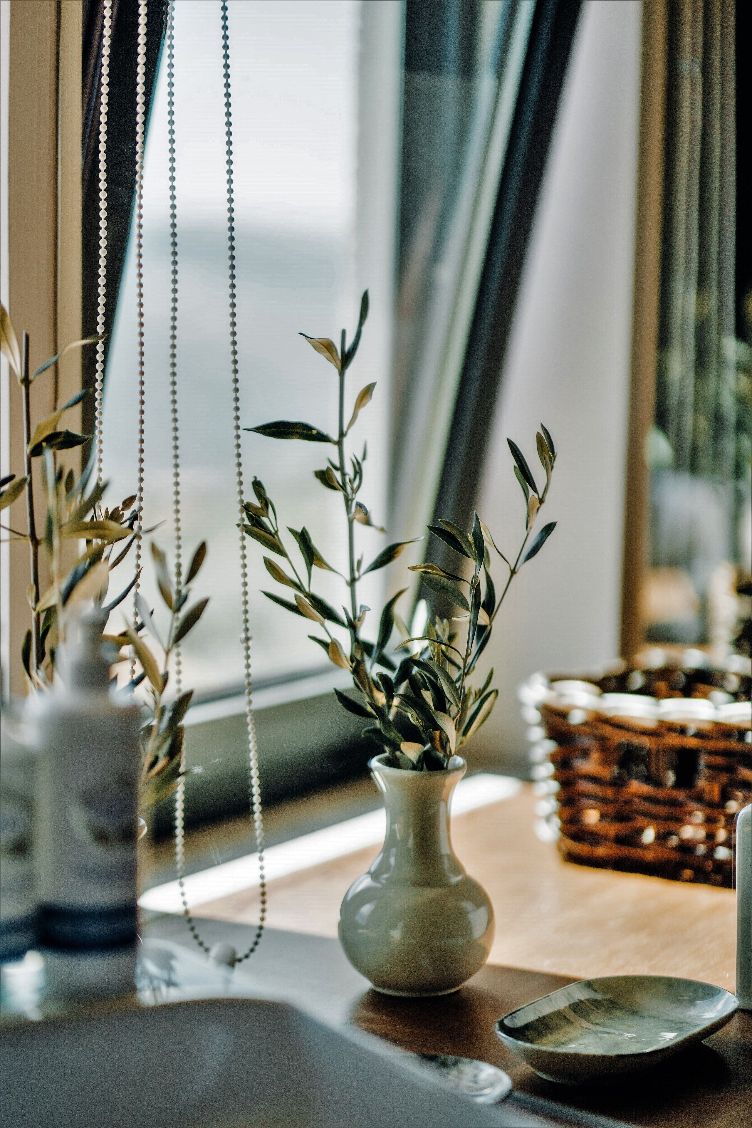 A room with a window, a vase with green leaves, a white ceramic bowl, and a wicker basket.