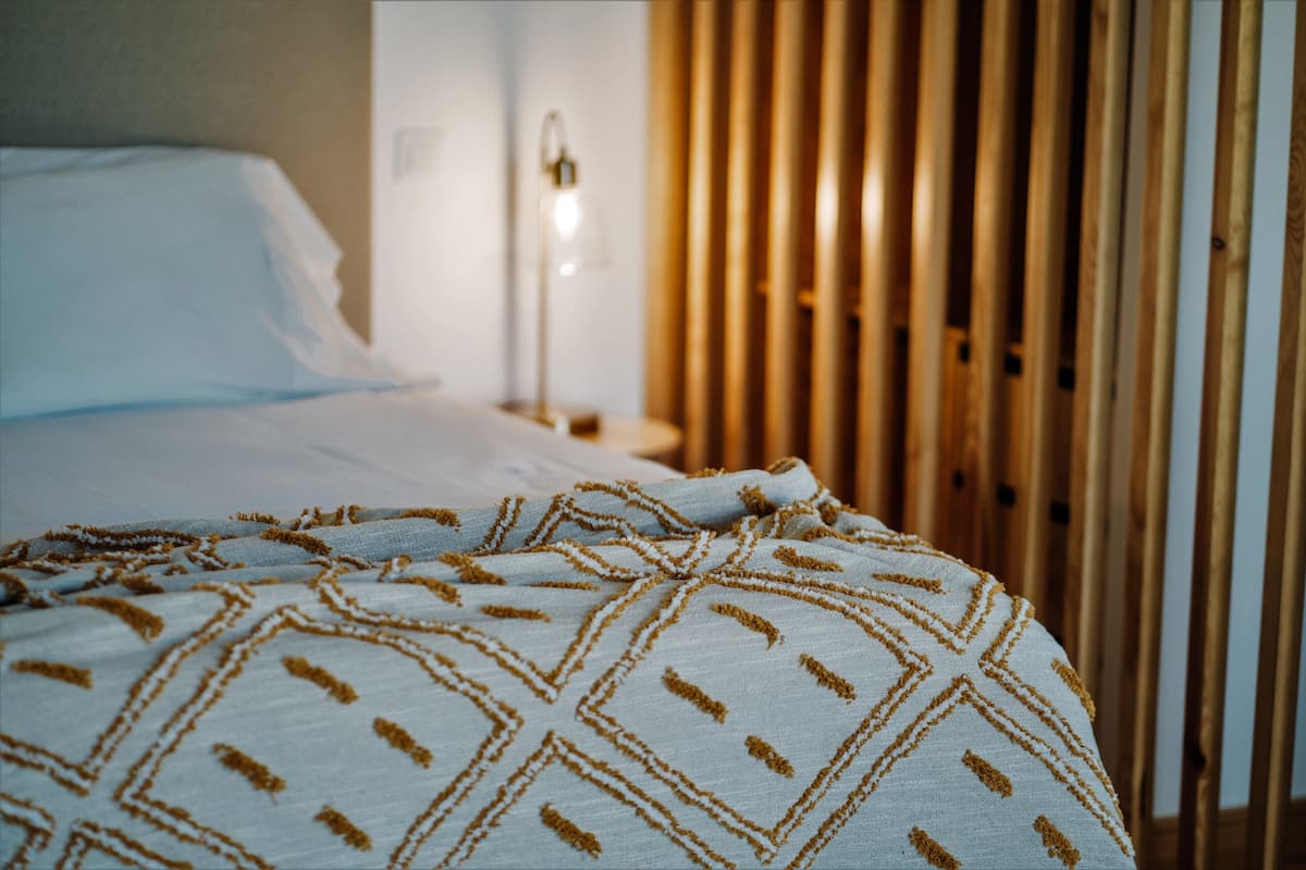 A bed with a patterned blanket, a bedside lamp, and wooden slats in the background.