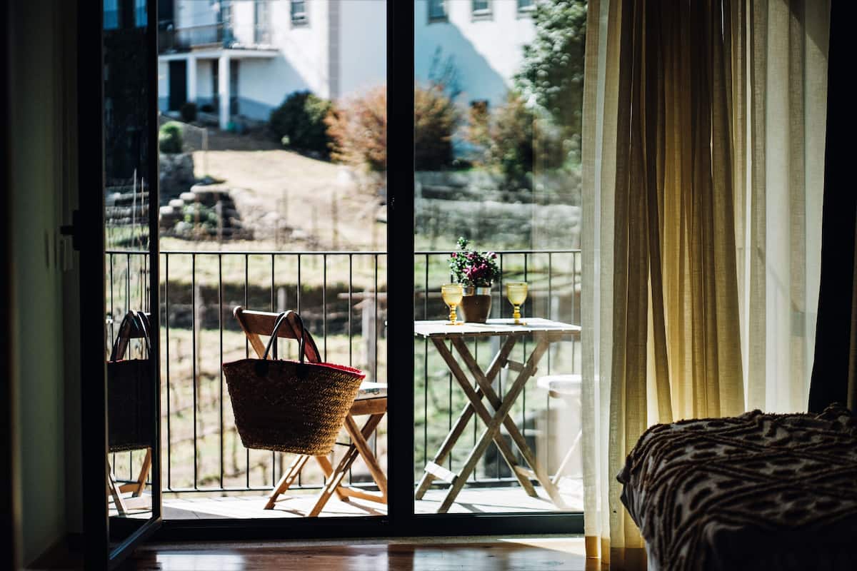 A balcony with a wicker chair, a small table with a vase and two glasses, and a view of a building and trees.