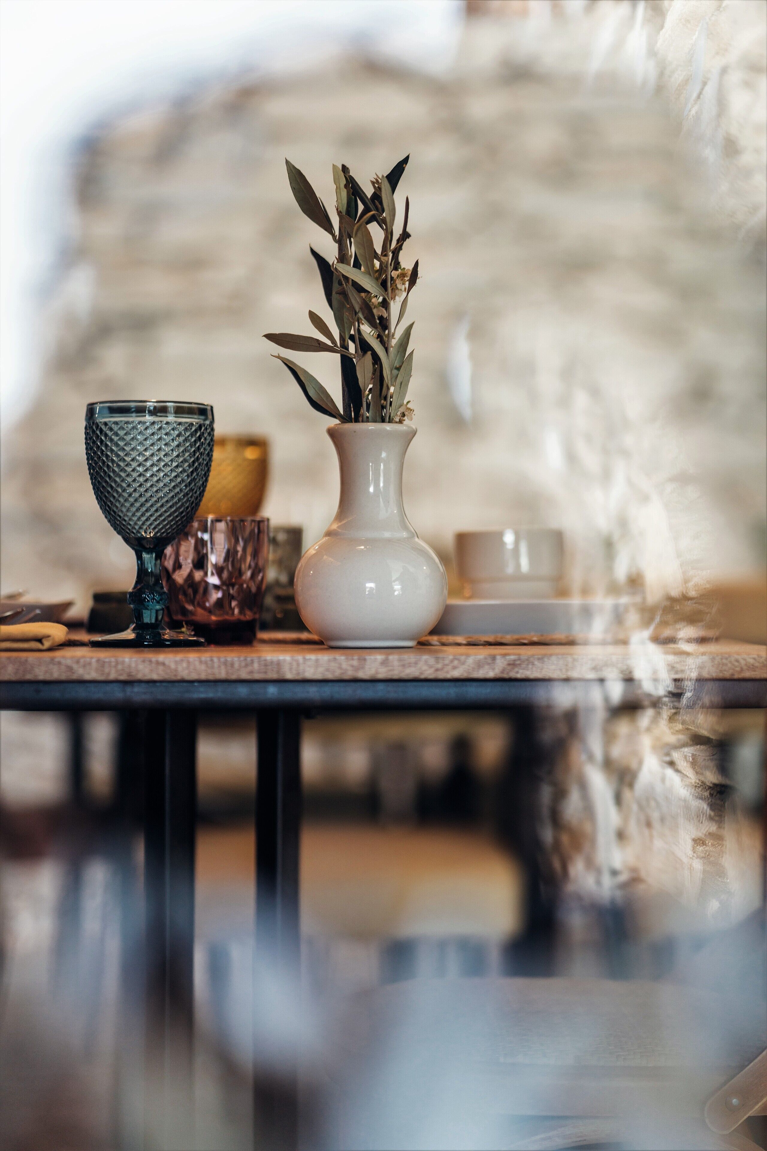 Una mesa con un jarrón blanco que contiene plantas secas, un cuenco de vidrio texturizado y una taza de vidrio transparente.
