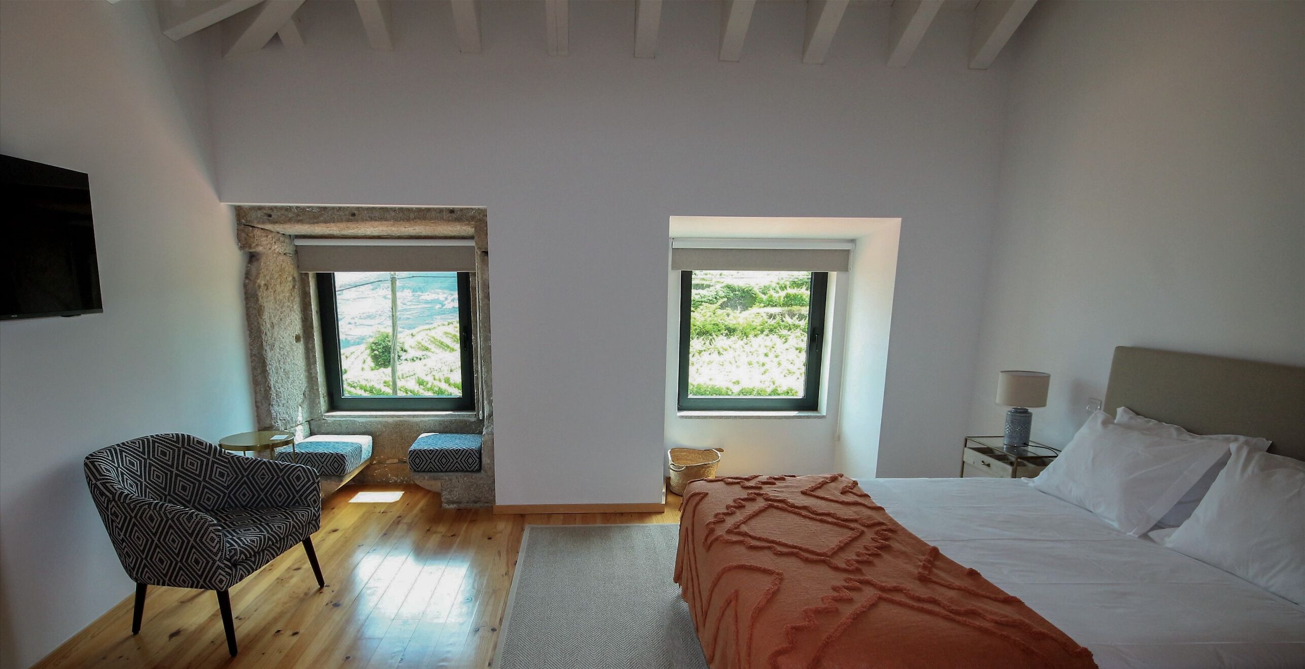 A bedroom with a bed, a chair, a small table, and a window with a view of greenery.