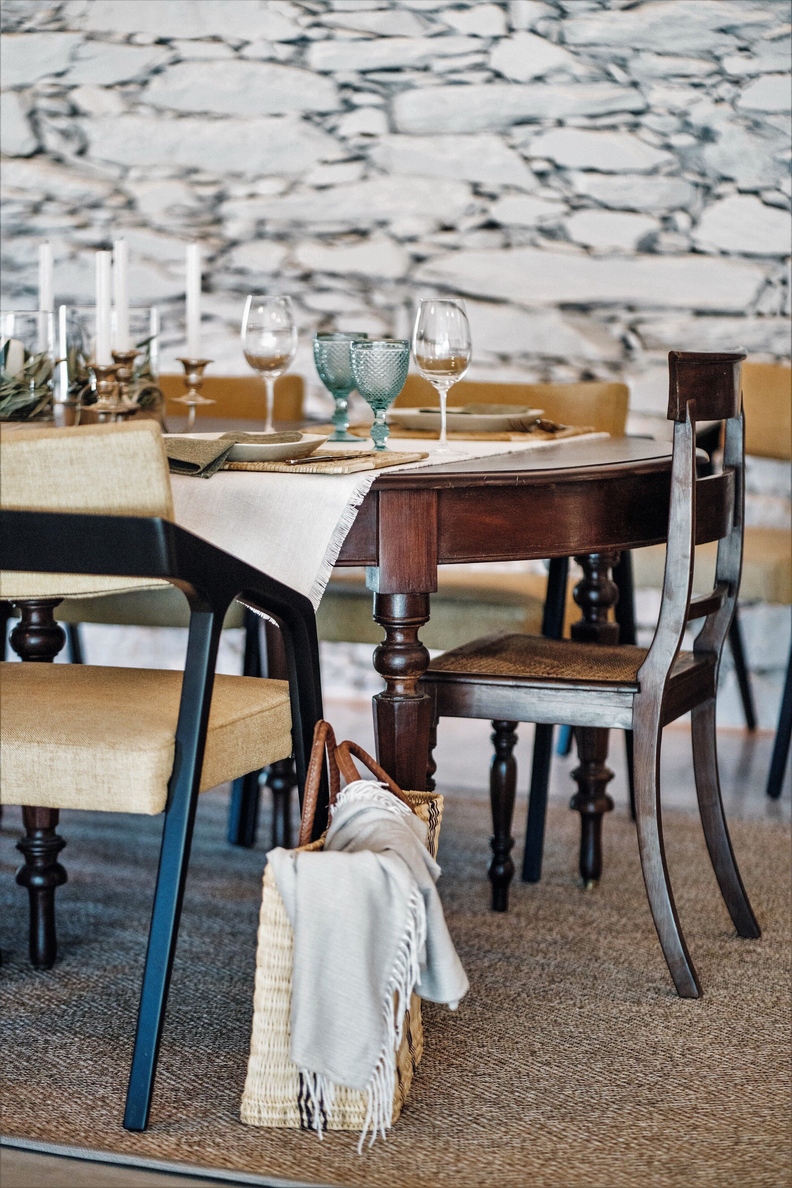 A dining area with a marble wall, wooden chairs, a table set with candles, glasses, and a woven basket.
