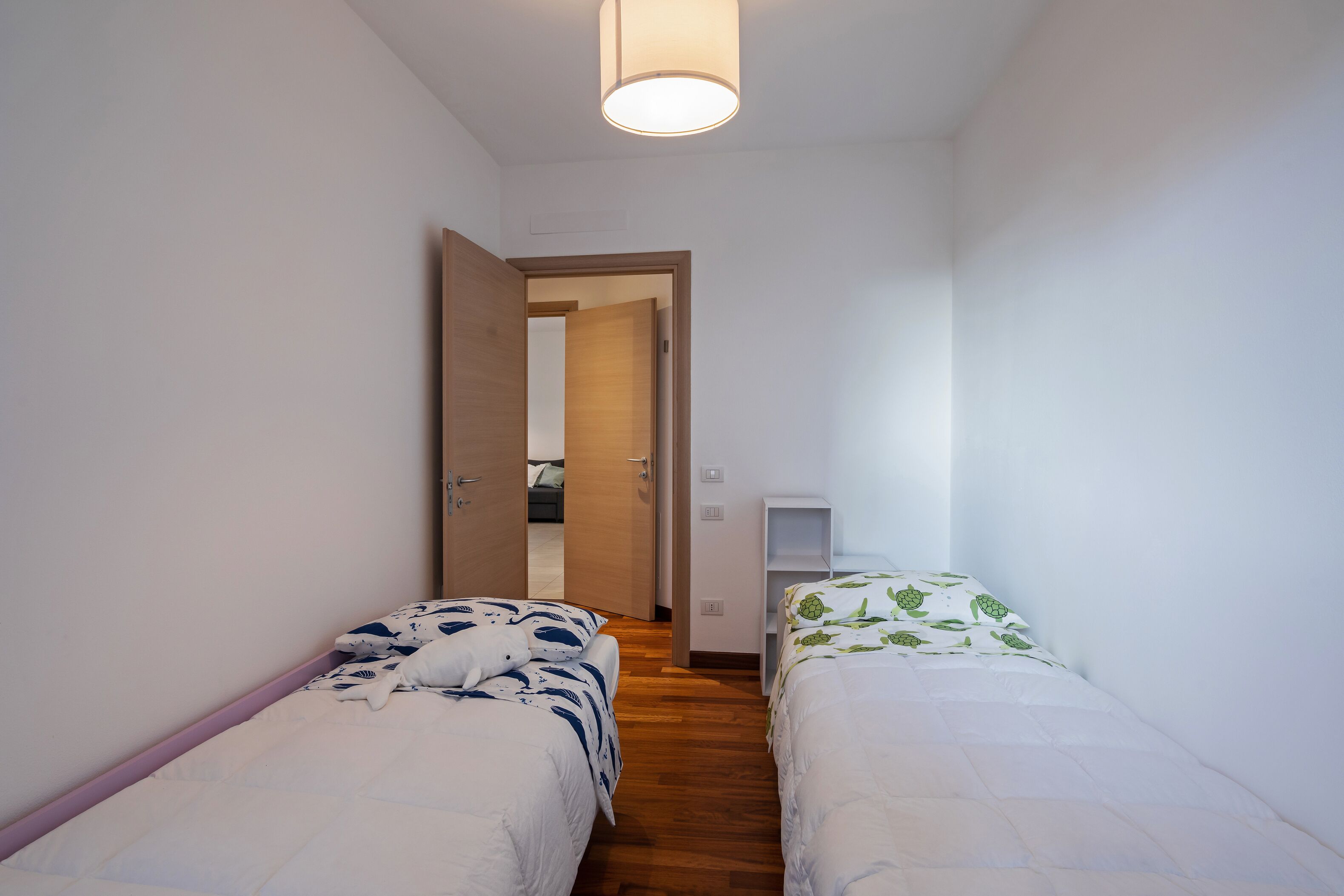 Habitación estrecha con dos camas, piso de madera y una puerta que da a otra habitación.