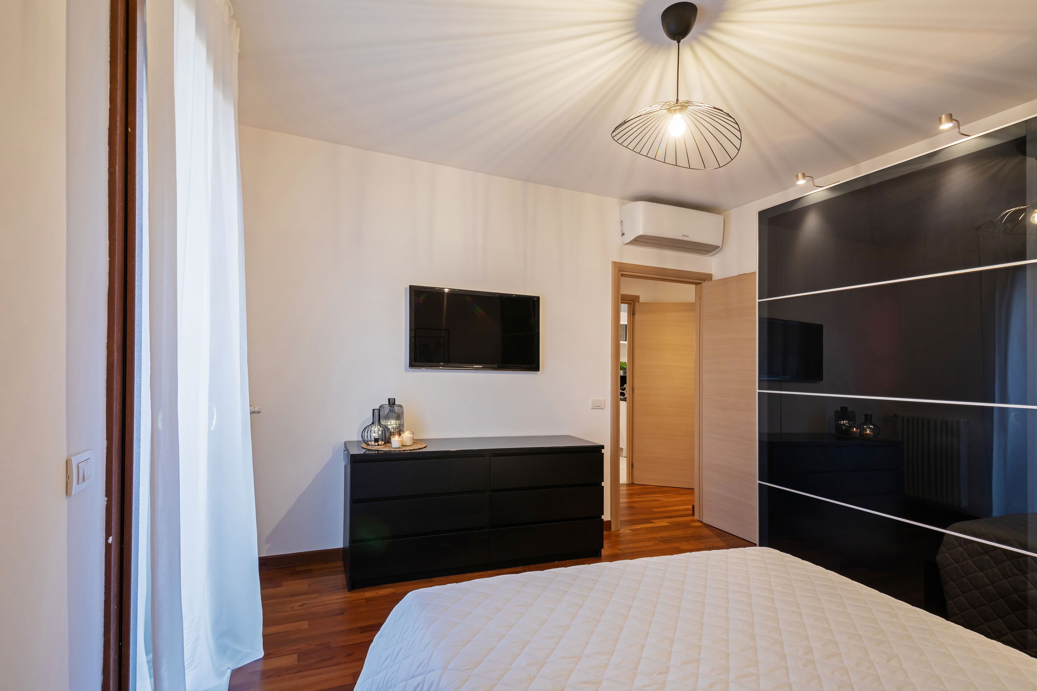 A modern bedroom with a large bed, a black dresser, a flat-screen TV, and a ceiling light fixture.