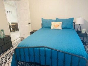 A bedroom with a blue bedspread, a metal headboard, a bedside table with a lamp, and a door leading to another room.