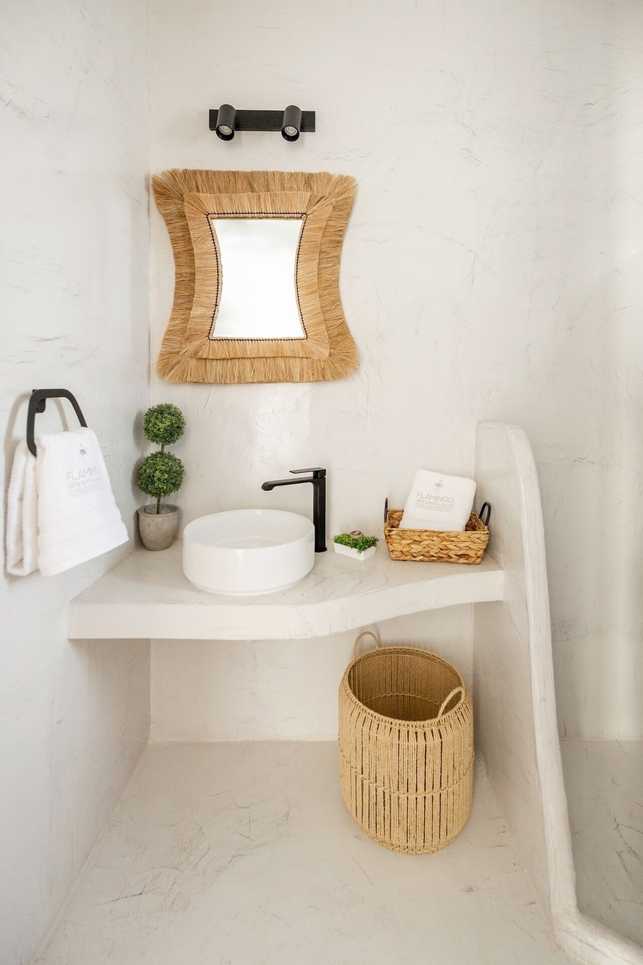 Casa de banho com lavatório branco, torneira preta e espelho com moldura de madeira. Há um porta-toalhas com toalha branca, uma pequena planta em vaso e uma cesta de vime com um saco.