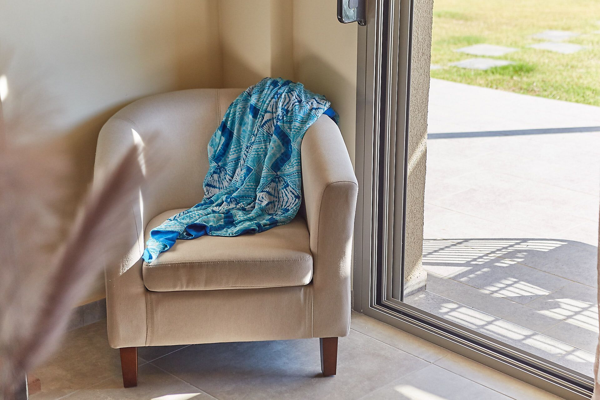 A beige armchair with a blue patterned throw blanket, positioned near a glass door leading outside.