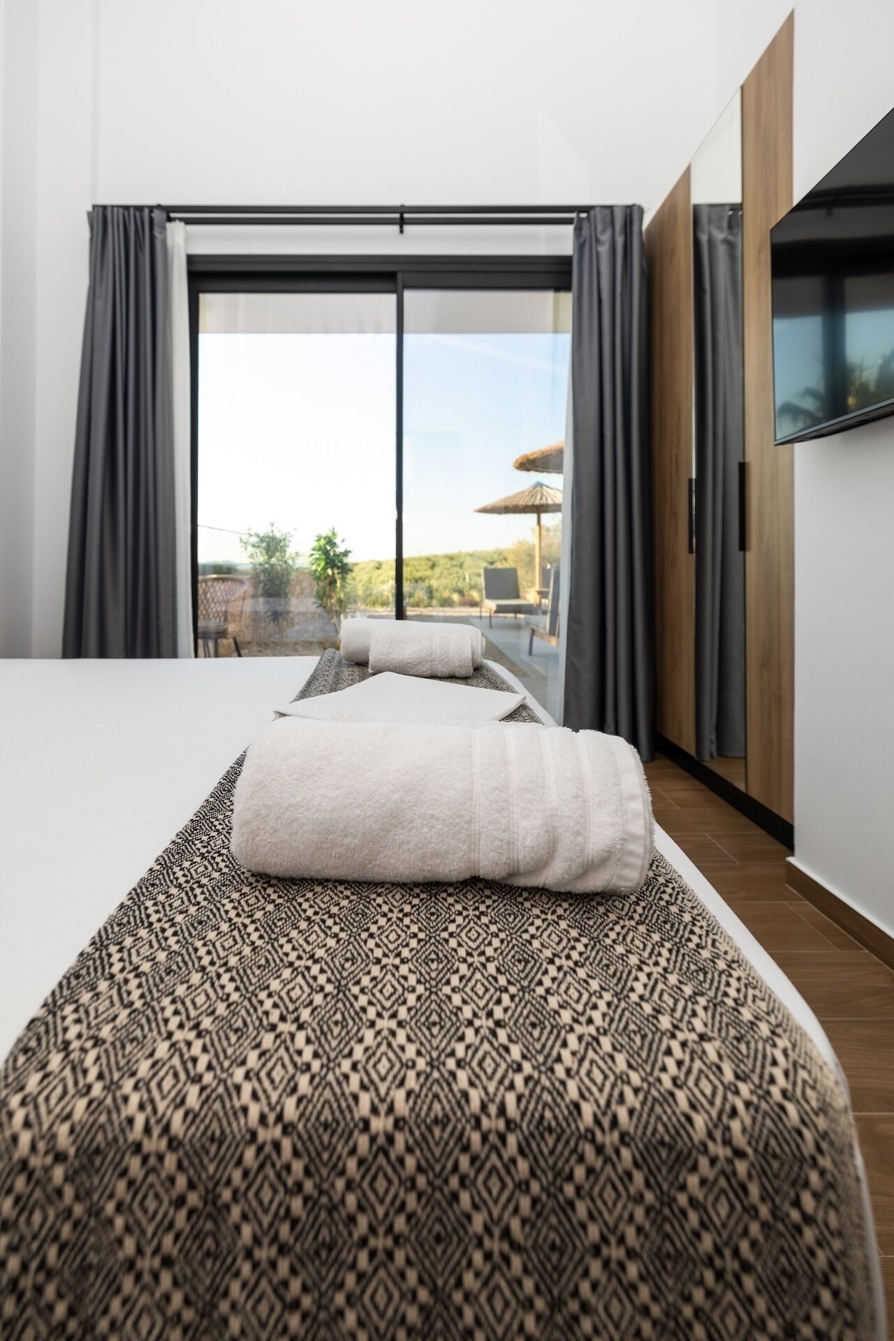 A modern hotel room with a large bed, a patterned bedspread, a view of the outdoors, and a flat-screen TV mounted on the wall.
