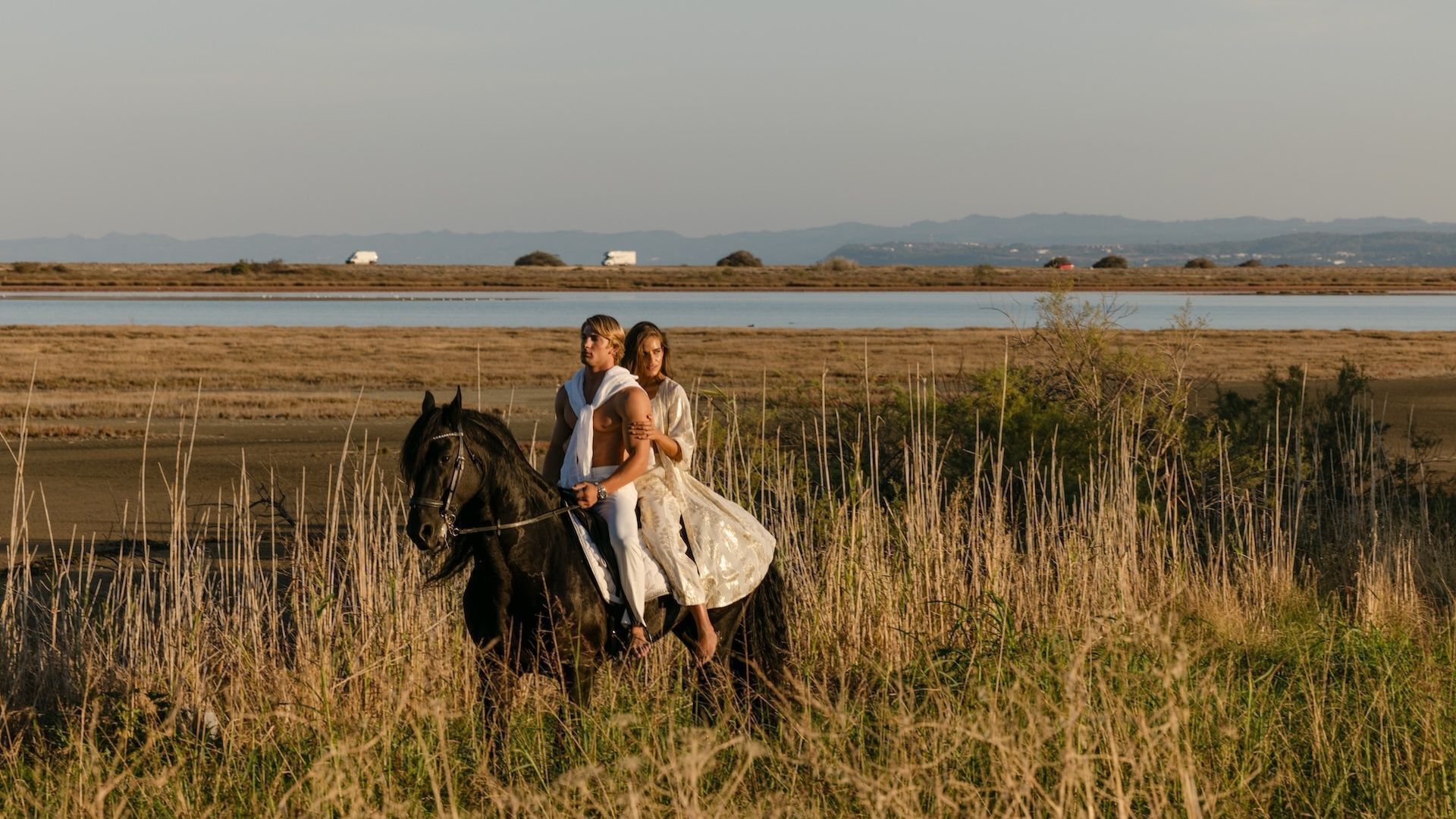 A horse and two people in a field with a body of water and distant hills in the background.