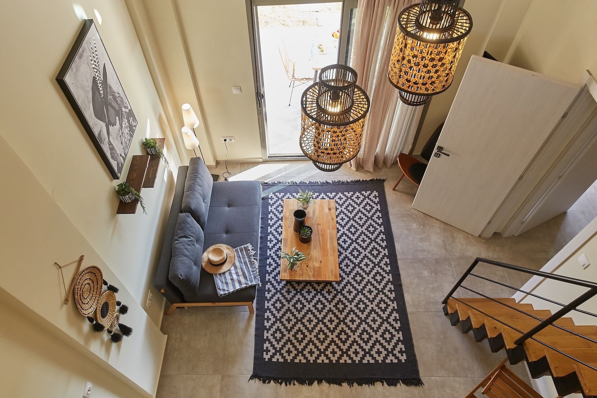 Ein modernes Wohnzimmer mit grauem Sofa, einem Holztisch und einem gemusterten Teppich. Zwei hängende Korblampen, ein gerahmtes Bild an der Wand und eine Treppe, die zu einer anderen Ebene führen.