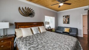 Een slaapkamer met een houten plafond, een bed met een beddegoed met dessin, een nachtkastje met een lamp, een kunstwerk aan de wand, een raam met jaloezieën, een grijze bank en een schilderij.