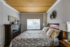 Une chambre avec un plafond en bois, un lit recouvert d’une couvre-lit à motifs, une table de chevet, une commode et un élément décoratif mural.