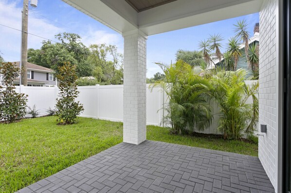 A covered patio area with a brick floor, surrounded by a white fence and lush greenery.