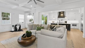 Un salon spacieux doté d’un grand ventilateur de plafond, d’un ensemble de canapés, d’une table basse et d’un coin cuisine avec des meubles blancs et une hotte noire.