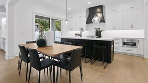 Une cuisine moderne avec des meubles blancs, une table à manger en bois, des chaises noires et des luminaires suspendus.