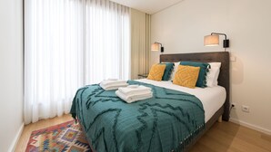 Ein ordentlich gemachtes Bett mit einer petrolfarbenen Tagesdecke und zwei Kissen, einem gemusterten Teppich und einer an der Wand befestigten Lampe.
