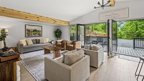 Ein geräumiges Wohnzimmer mit einem grauen Sofa, beigen Sesseln, einem Couchtisch und einem Holzbalken. Ein großes Fenster bietet Blick auf eine Terrasse und Bäume.