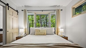 Ein Schlafzimmer mit einem großen Bett, zwei Nachttischen inklusive Lampen, einem Fenster mit Vorhängen und Blick auf Bäume.