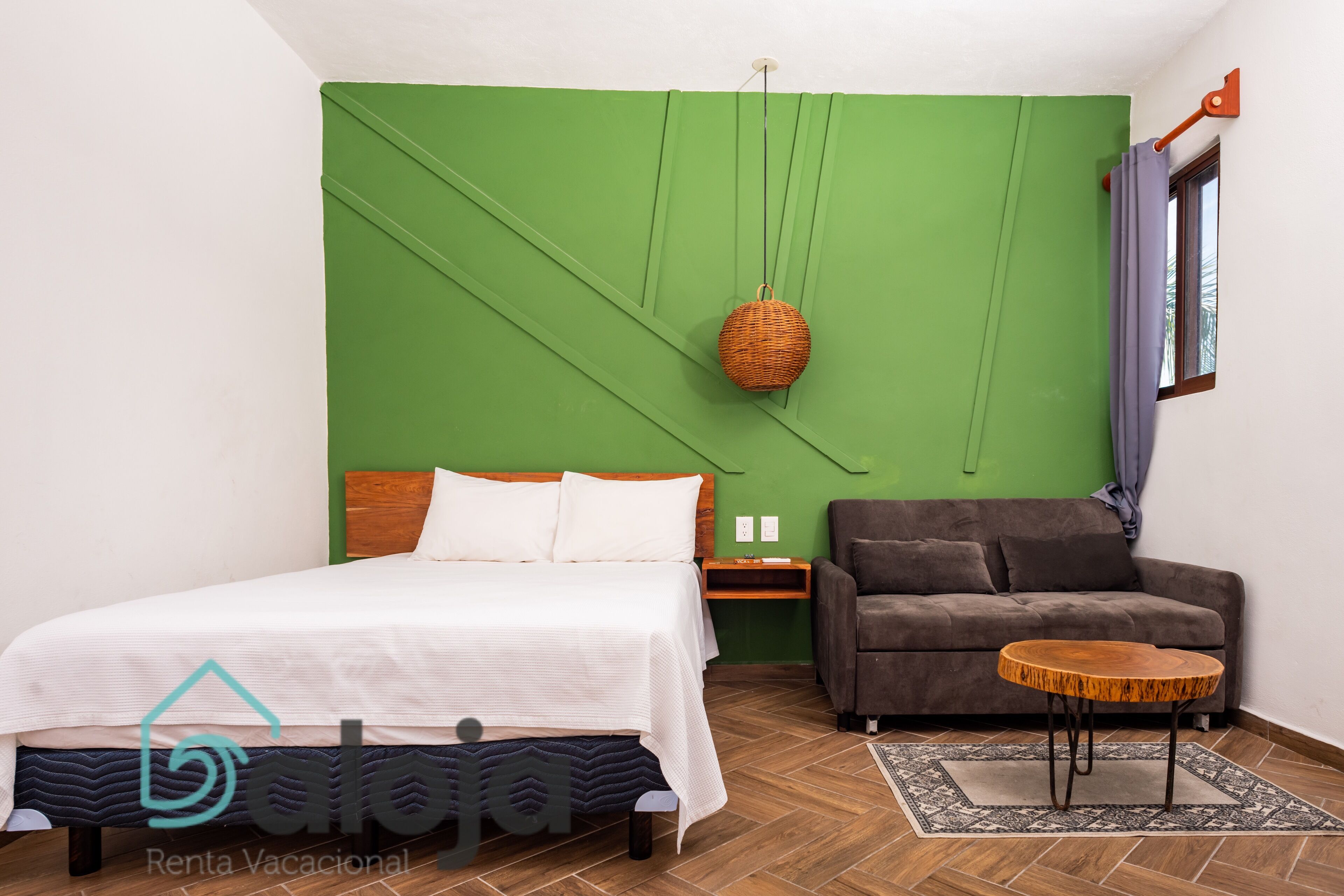 A bedroom with a bed, a green accent wall, a hanging basket, a sofa, and a small round table.