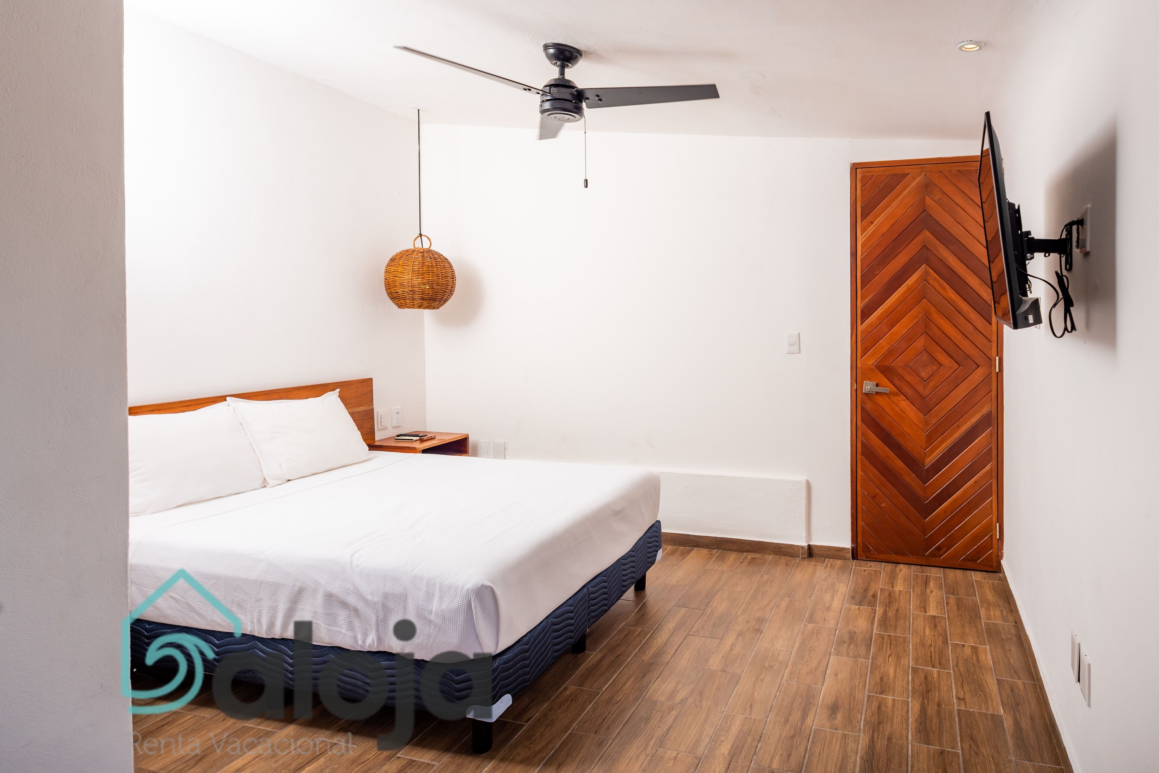 A bedroom with a bed, a ceiling fan, a wall-mounted TV, a wooden door, and a woven light fixture.