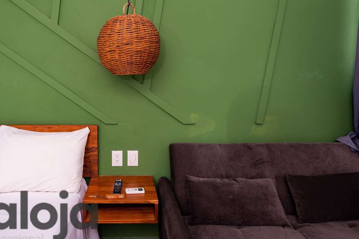 A room with a bed, a brown couch, a hanging wicker lamp, and a wooden nightstand with a remote control and a phone.