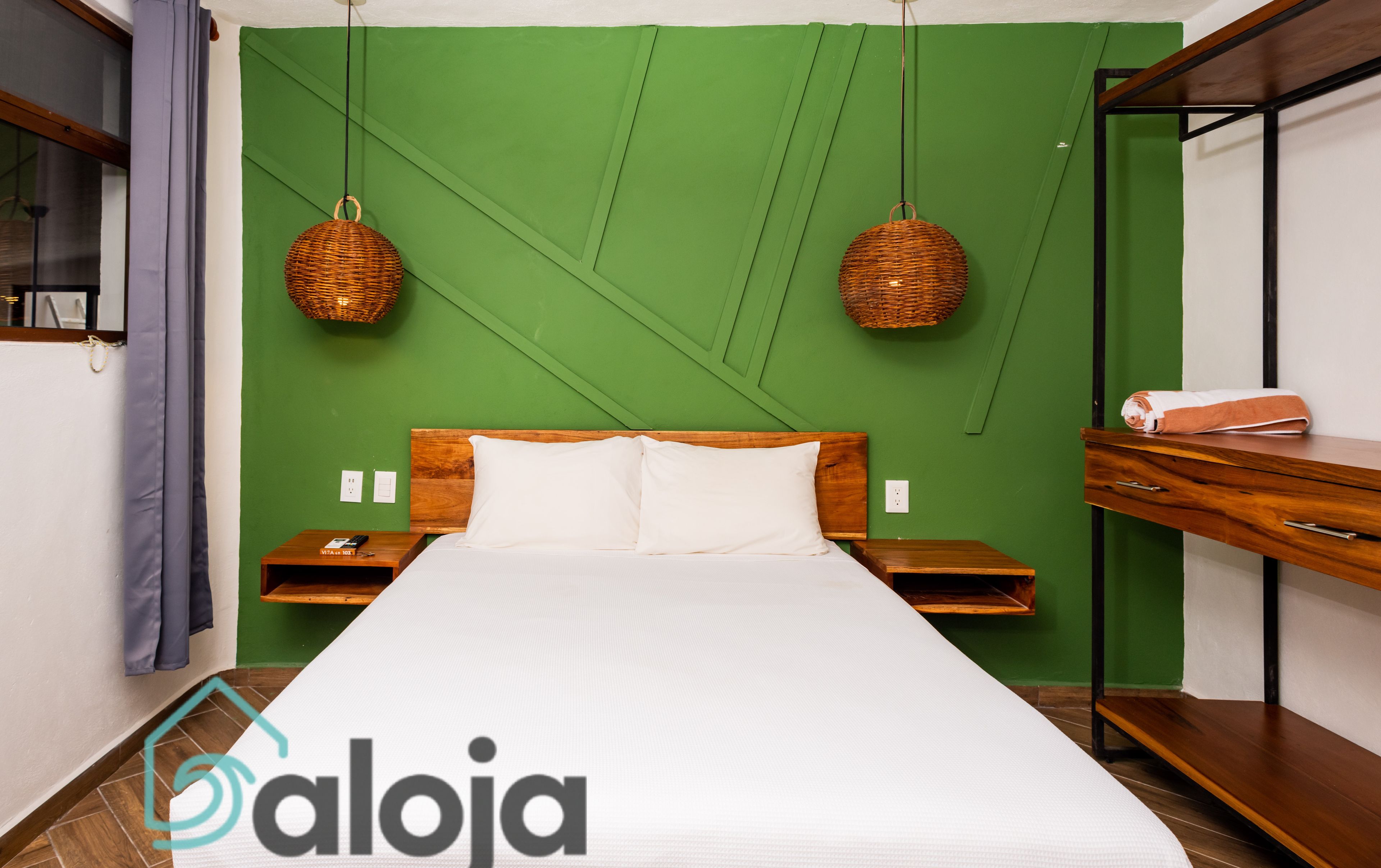 A neatly made bed in a room with green walls, two wicker wall hangings, a wooden nightstand, and a built-in bench with a towel.