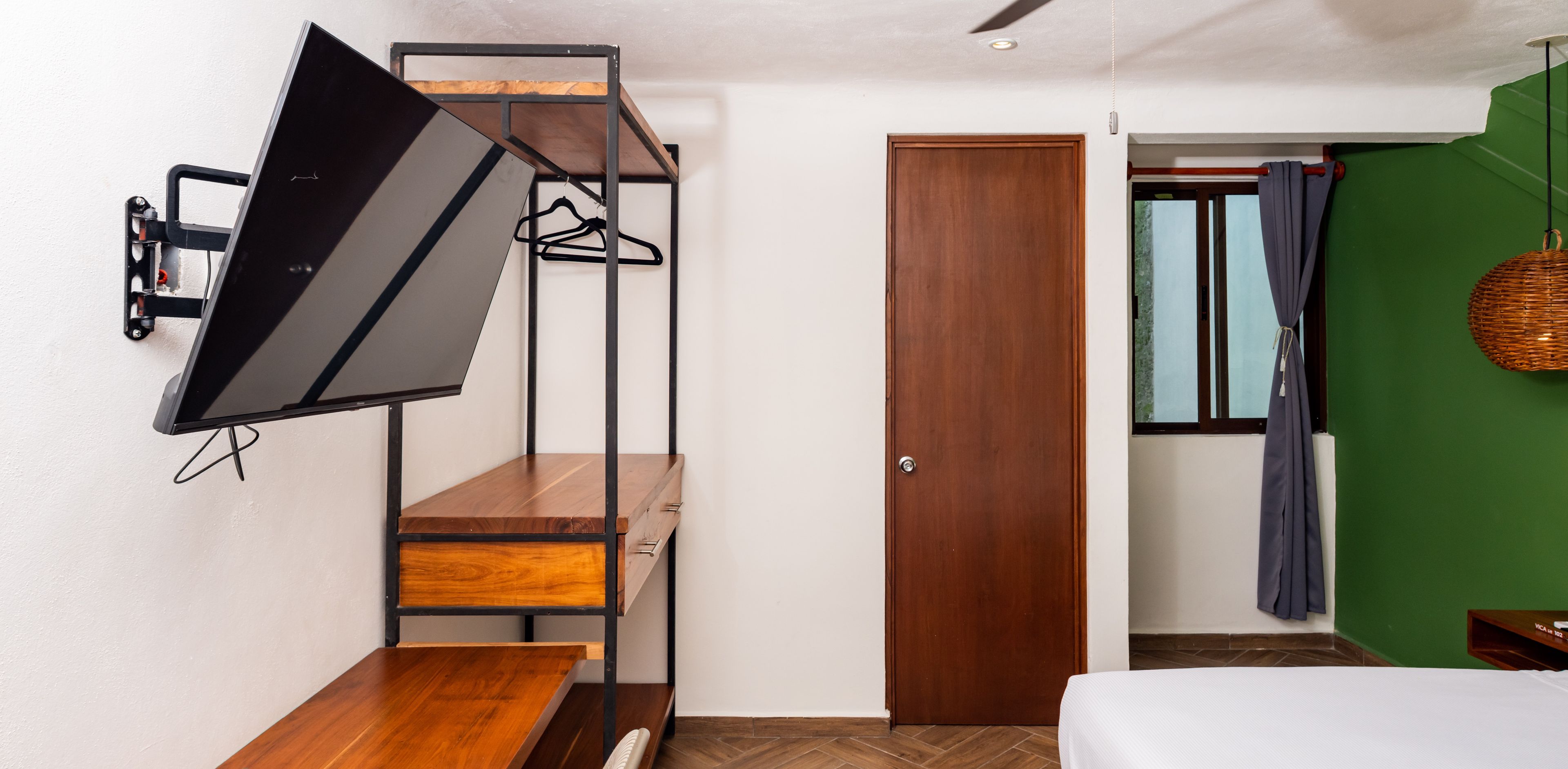 A compact room with a bed, a desk with a chair, a wall-mounted TV, a wardrobe, and a window with curtains.