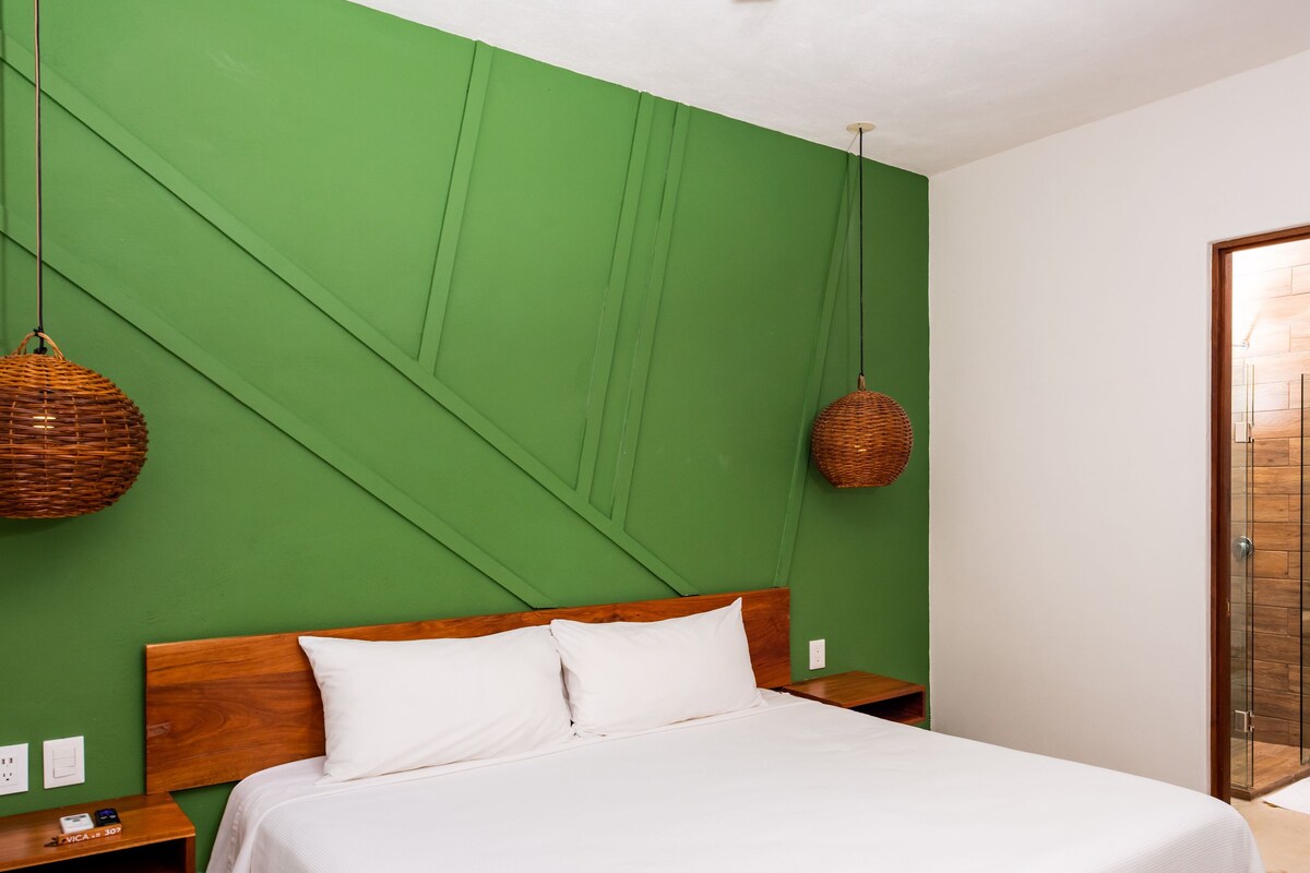 A bedroom with a bed, green walls, a wooden headboard, two wicker lamps, a wooden nightstand, and a bathroom visible through a glass door.