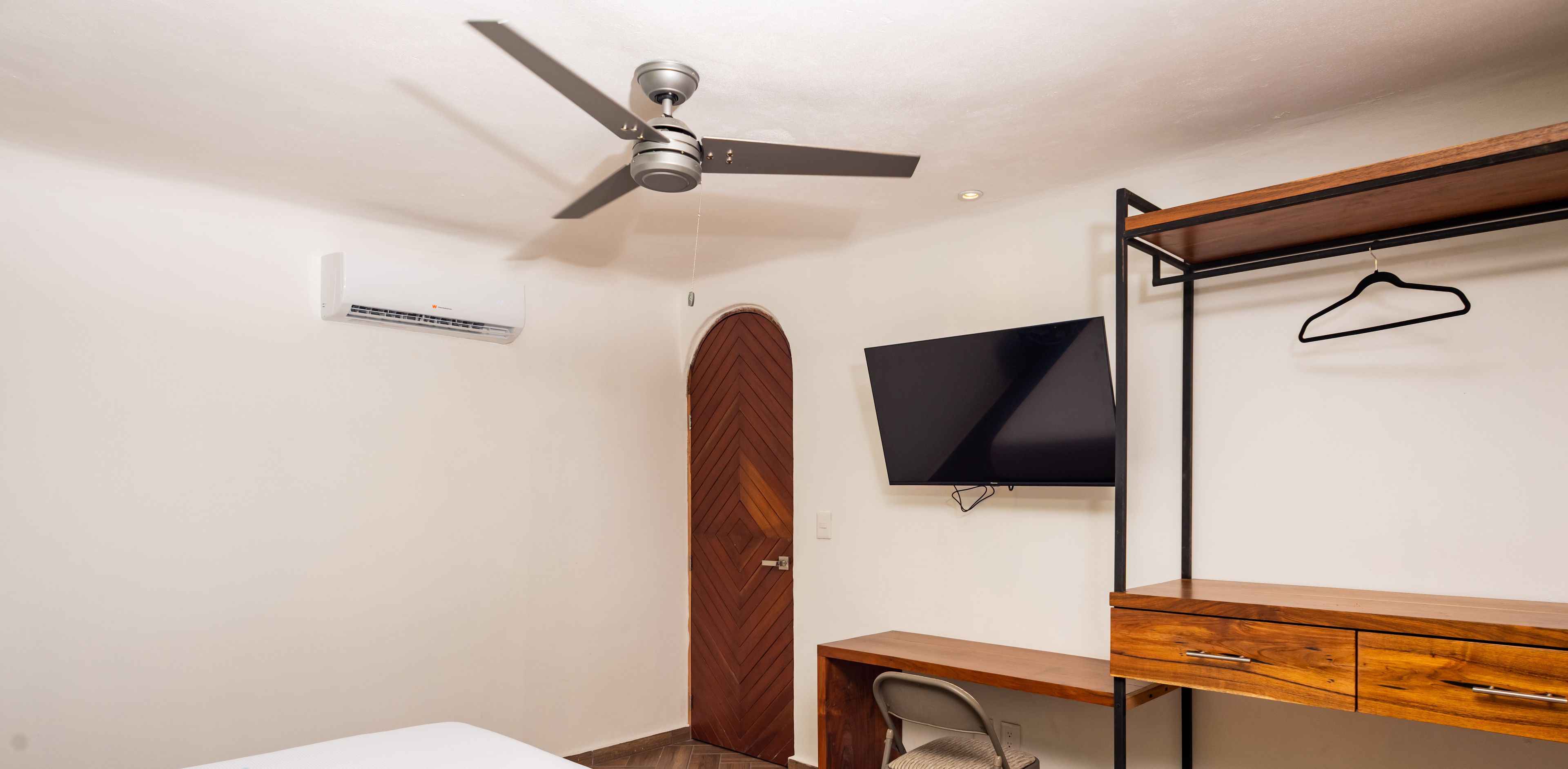 A hotel room with a bed, a desk with a chair, a TV, a wardrobe, and a ceiling fan.