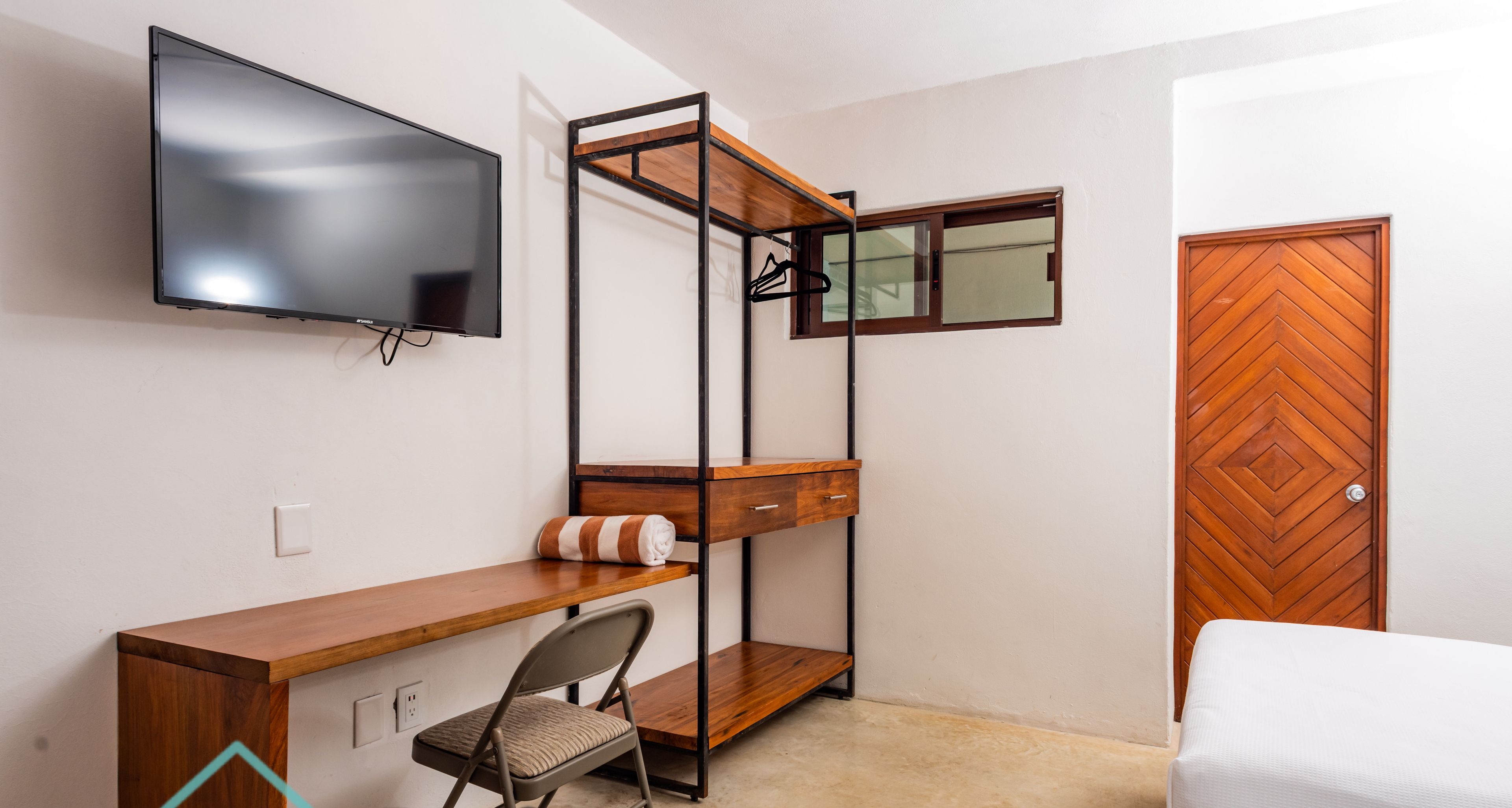 A compact room with a wooden desk, a chair, a bed, a TV mounted on the wall, and a wardrobe.