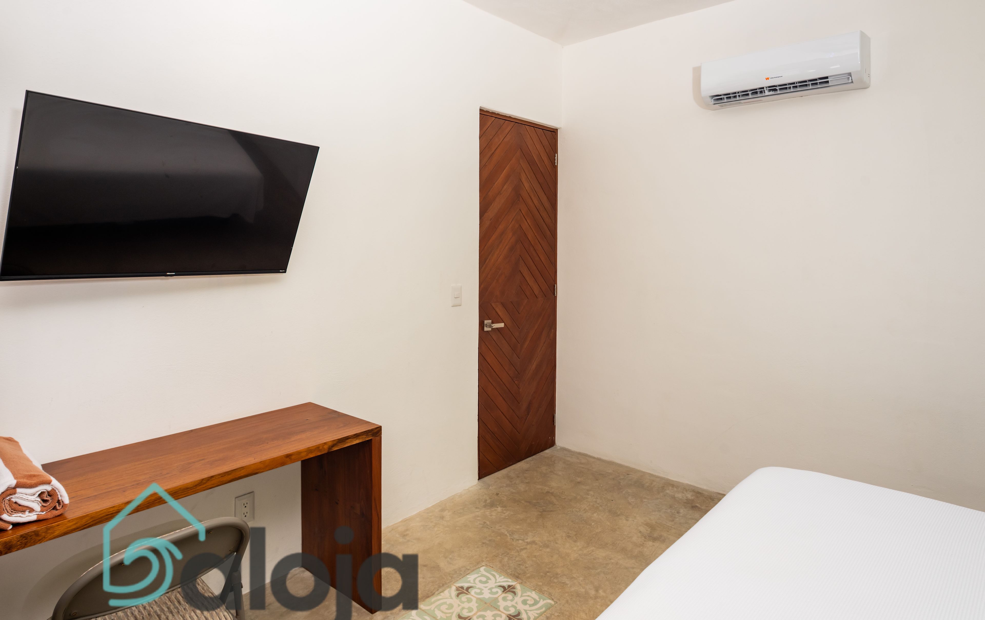 A room with a flat-screen TV mounted on the wall, a wooden door, an air conditioning unit, a small wooden desk with a chair, and a bed with a white mattress.