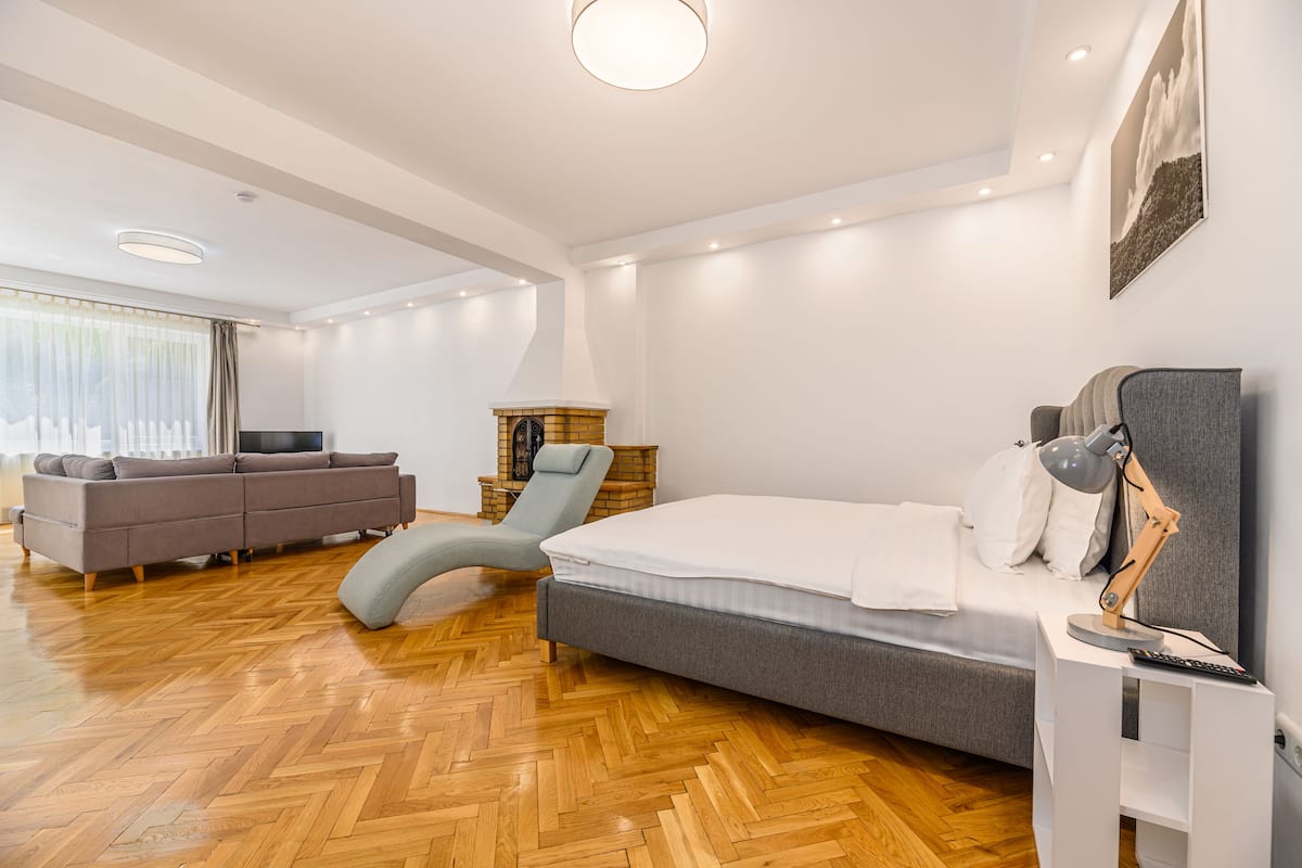 Un dormitorio moderno con suelo de parqué de madera, una cama y un sofá grises, un sillón azul y un pequeño armario de madera.