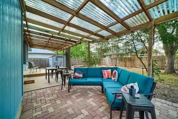 Relax on the patio with a blue sofa, round table, and stool, surrounded by trees and a wooden fence.