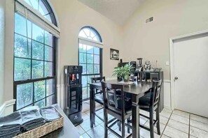 Enjoy meals in this dining area with a table, chairs, and a coffee machine, surrounded by a potted plant and a view of the trees outside.