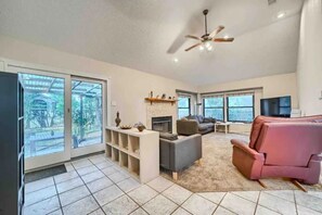 Relax in this spacious living room with a fireplace, sofa, and armchair, leading to a patio with a view of the surrounding greenery.