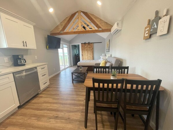 A compact kitchen with white cabinets, a wooden dining table with chairs, and a lofted ceiling with exposed beams.