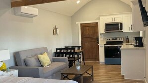 A spacious room with a loft bed, a gray sofa, a wooden dining table, and a kitchen area with white cabinets and appliances. - Modern wine country barn house (Temecula)