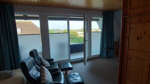 Ein Zimmer mit Holzboden, einem Sofa, einem Hocker und einer Glastür zum Balkon.