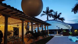 An outdoor dining area with string lights, a large spherical decoration, and a covered seating area with a pool in the background. - Hotel Hacienda el Novillero (Fusagasugá)