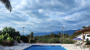 A swimming pool with lounge chairs and umbrellas, surrounded by greenery and mountains in the background. - Hotel Hacienda el Novillero (Fusagasugá)