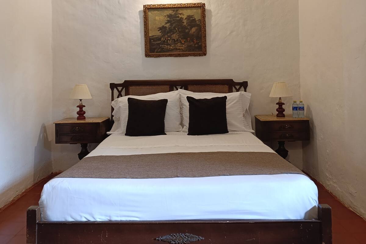 Une chambre avec un lit en bois, deux tables de chevet avec des lampes, un tableau au mur et un sol carrelé.