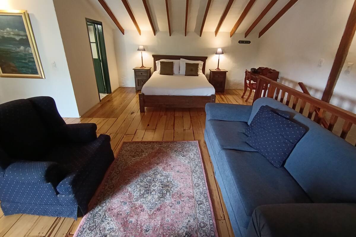 Une chambre avec un grand lit, deux tables de chevet avec des lampes, un canapé bleu, un tapis à motifs, des poutres en bois au plafond et un tableau encadré au mur.