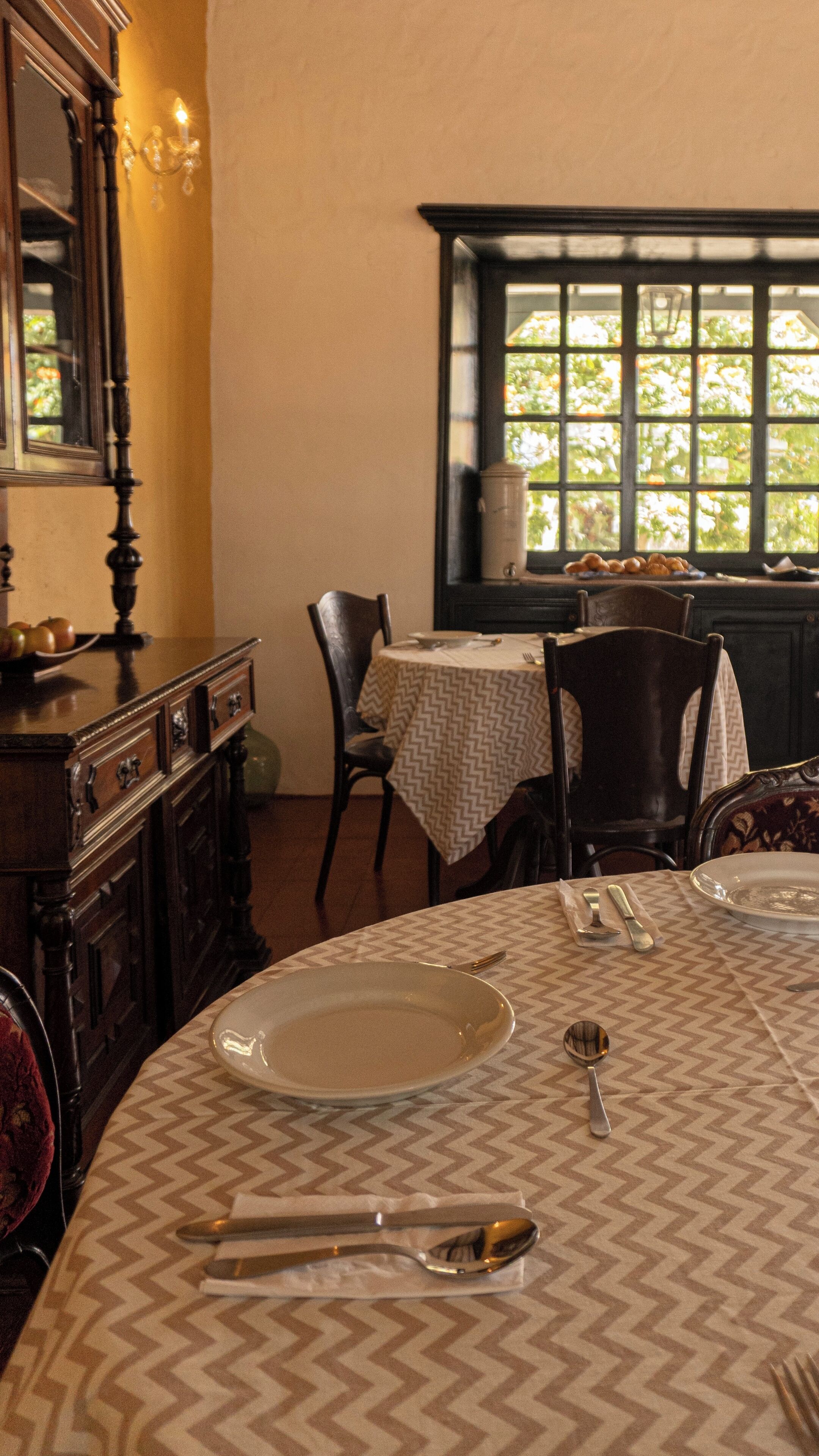 A dining area with a table set for two, a mirror, and a window with a view of greenery.